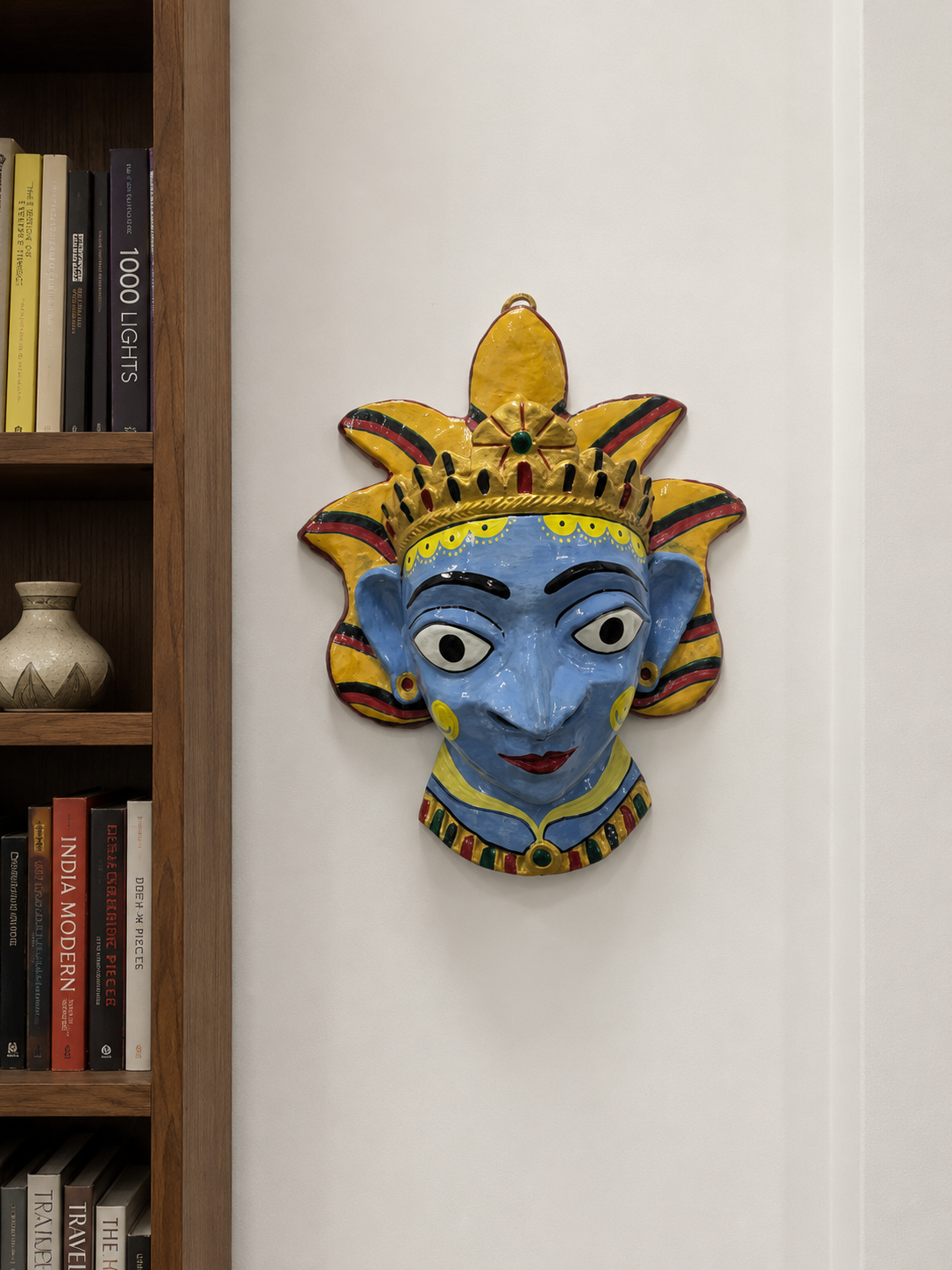 A hand-painted papier mache wall mask of a stylized face with a crown, mounted on a white wall.