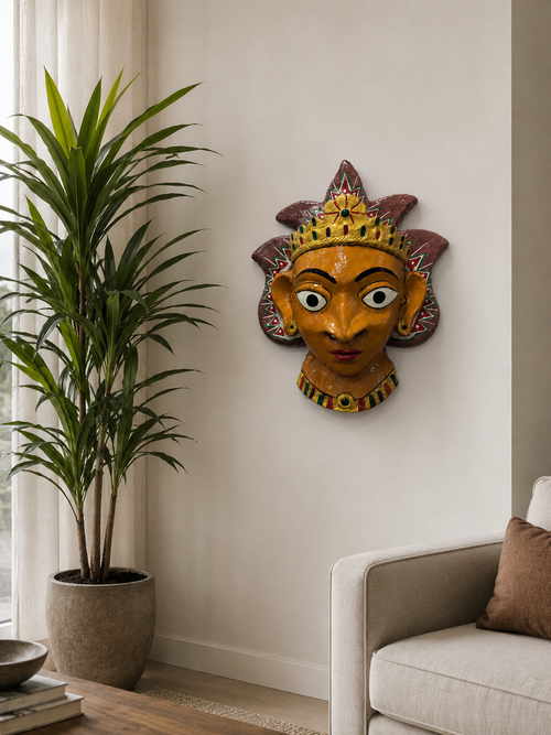 A decorative papier mache wall mask with a golden-brown face, red and purple headdress, and green accents, mounted on a beige wall in a modern living room setting next to a potted plant and a sofa.

Details