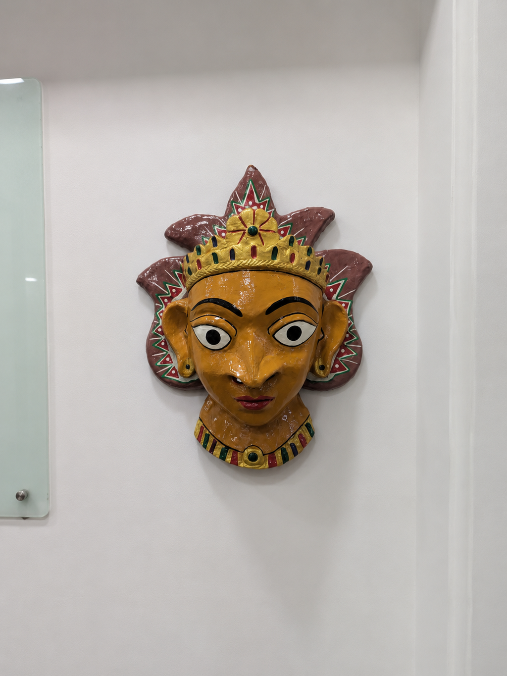 A hand-painted papier mache wall mask of a stylized face with a crown, mounted on a white wall.